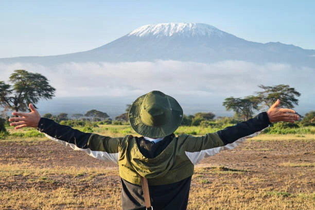 Person standing with arms outstretched facing Mount Kilimanjaro rising above the clouds in the distance.